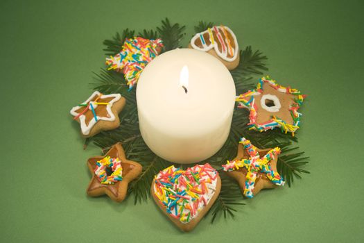White pillar candle on fir branches surrounded by decorated gingerbread cookies on green background