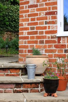 The corner of a set of stone and brick constructed steps leading to and from a garden patio area with various clay, terracotta, tin and plastic pots on the steps. Various seasonal plants grow in the pots.