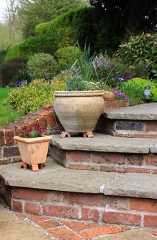 The corner of a set of stone and brick constructed steps leading to and from a garden patio area with two terracotta pots on the steps. Various seasonal plants grow in the pots. Set against a lush green garden background.