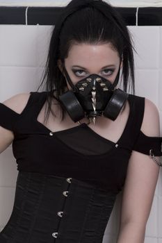 teenage girl wearing gothclothes with black hair and a gas mask agaisn't a ceramic wall