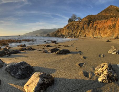 Steps in the sand at Big Sur, California with beautiful sand, cliffs, sky and ocean in the background and rocks in the foreground.
