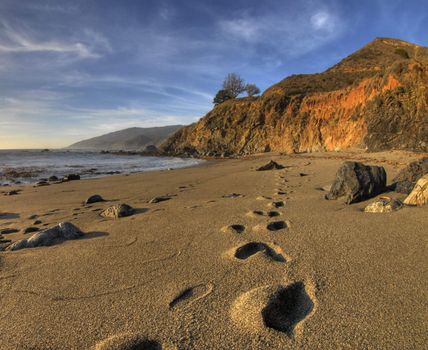 Footprints heading off into the distance on the beach at Big Sur, California with the water, cliffs and a beautiful sky in the background