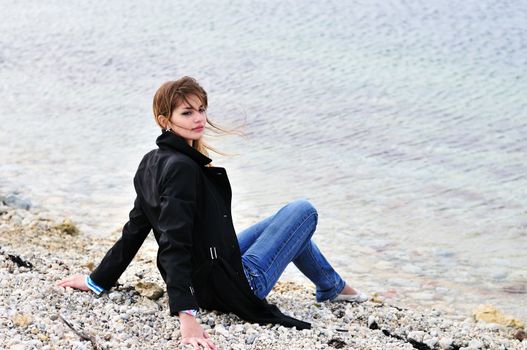  lovely teen girl sitting on the pebble close to sea in spring time
