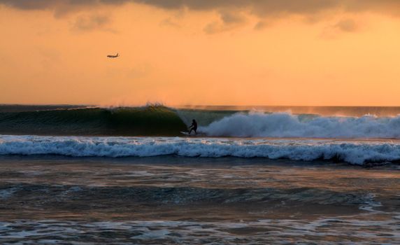 Surfer rides waves at sunset, airplane landing in the background