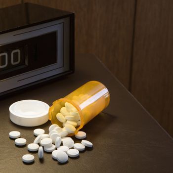 Open medicine bottle with pills spilling out onto table with clock in background.