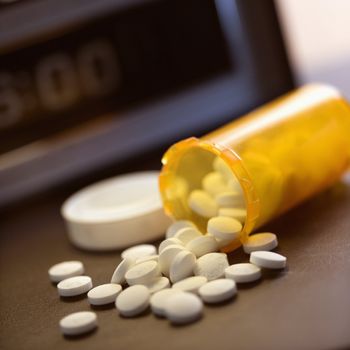 Open medicine bottle with pills spilling out onto table with clock in background.