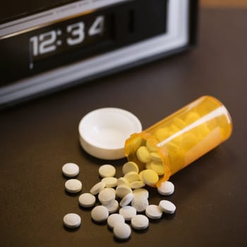 Open medicine bottle with pills spilling out onto table with clock in background.