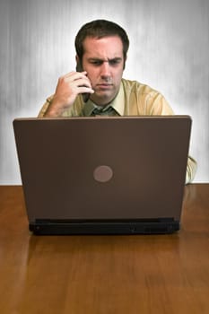 A man working from home with his cell phone and laptop. He has an upset or serious look on his face.