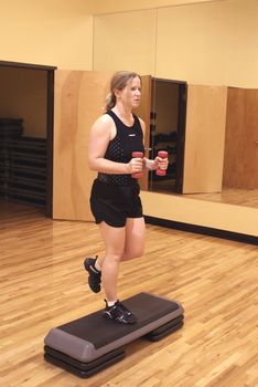 Woman working out in a gym using hand weights and a step.              