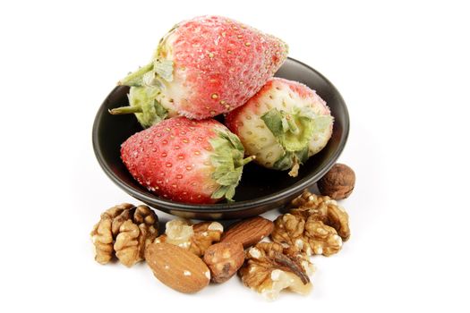 Red ripe frozen strawberries in a small black bowl with mixed nuts on a reflective white background