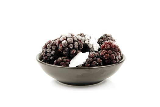 Ripe frozen blackberries in a small black bowl on a reflective white background
