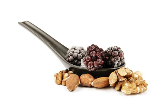 Ripe frozen blackberries on a small black spoon with mixed nuts on a reflective white background