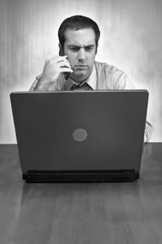 A man working from home with his cell phone and laptop in black and white. He has an upset or serious look on his face.