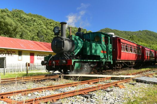A green locomotive hauls red carriages past a period hotel on the West Coast Wilderness Railway, as it travels from Queenstown to Strahan. Tasmania, Australia