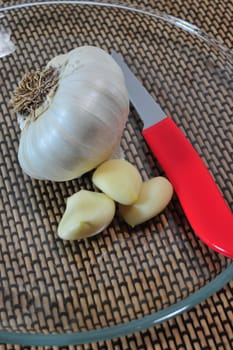 Whole garlic and pealed cloves on  clear glass plate with a red handled knife.