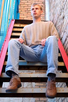 Young stylish man with blonde hair sit on stairs near brick wall.