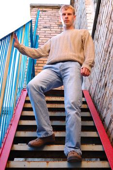 Young stylish man with blonde hair stay on stairs near brick wall.