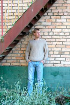 Young stylish man with blonde hair stay under stairs near brick wall.