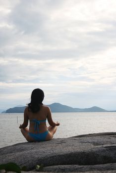 A girl meditates on a rock by the beach.