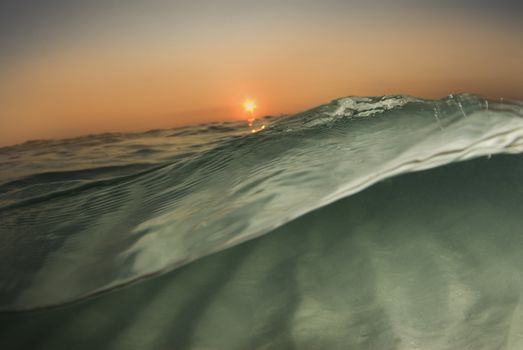 A wave passes by the camera half in, half out of the water while the sun sets on the horizon and the ripples of the ocean floor reflect off the surface.