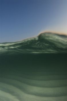 Curl of a passing wave on an underwater/overwater shot image with the blue sky and color from the setting sun rim lighting the wave.