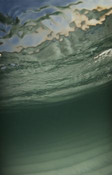 Underwater view of the sea floor with the ocean surface and the bright blue sky and setting sun showing through the surface.