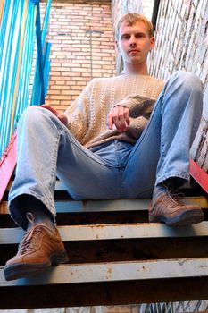 Young stylish man with blonde hair sit on stairs near brick wall.