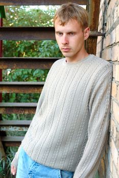 Young stylish man with blonde hair stay under stairs near brick wall.