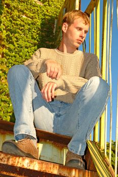 Young stylish blonde man sit on stairs near overgrown with vine wall.