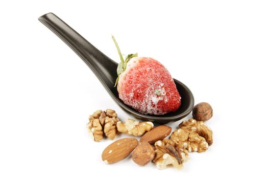 Red ripe frozen strawberry on a small black spoon with mixed nuts on a reflective white background