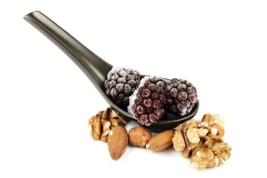 Ripe frozen blackberries on a small black spoon with mixed nuts on a reflective white background