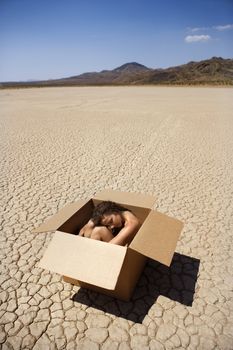 Pretty nude young woman sitting in box in cracked desert landscape in California.