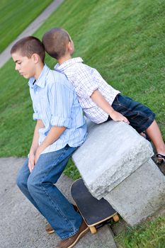 Two brothers sitting back to back on a bench in the park looking very bored.  