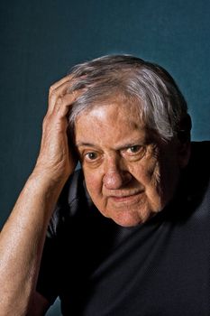 Dramatic portrait of a senior man with his hand on the side of his face and in his hair wearing a black shirt isolated on gray/blue