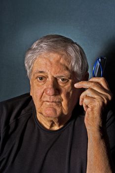 Dramatic side portrait of a senior man with a pair of glasses in his hand next to his face wearing a black shirt isolated on gray/blue
