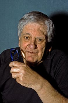 Dramatic portrait of a senior man with a pair of blue glasses in his hand wearing a black shirt isolated on gray/blue