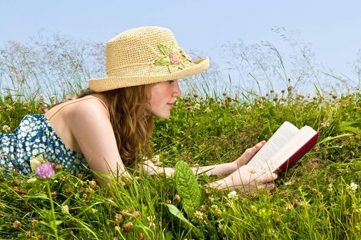 Young teenage girl reading book in summer meadow with straw hat