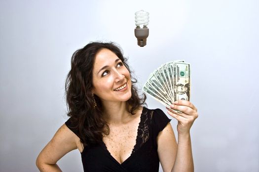 Woman waving money and looking up. Having an environmentally friendly idea with an energy saving fluorescent light bulb floating above her head, on a white background