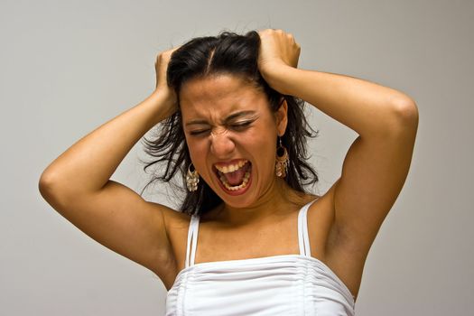 A young beautiful latina woman screeming of frustration and pulling her hair with both hands wearing a white shirt, isolated on white