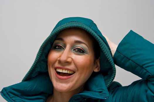 Beautiful smiling young woman wearing a green winter coat with the hood over her head and her hand behind her head, isolated on white