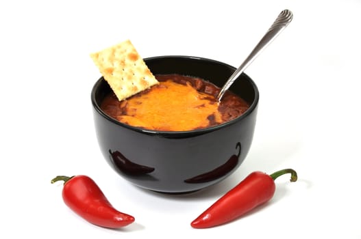 Bowl of chili with melted cheese, cracker, red cayenne peppers, and spoon.  Isolated on white background.