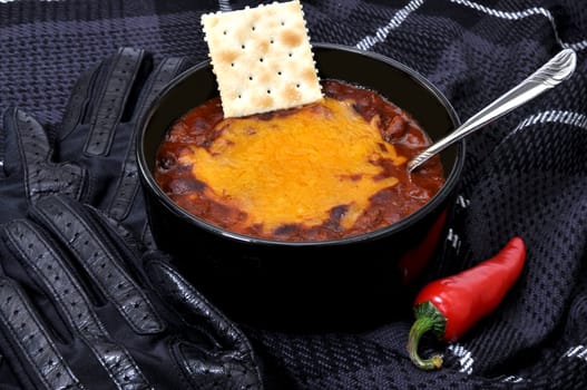 Bowl of chili with melted cheese, cracker and red cayenne pepper.  Pair of gloves and scarf in background.