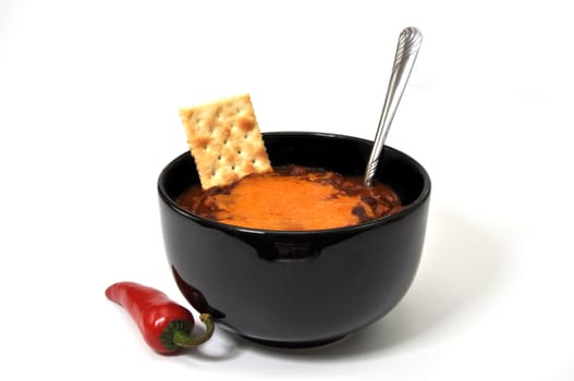 Bowl of chili with melted cheese, cracker, red cayenne pepper, and spoon.  Isolated on white background.
