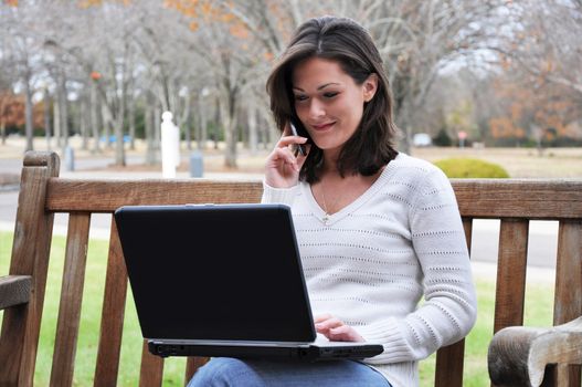 Young woman sitting on bench in park talking on cell phone and using laptop.