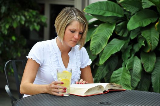 Blond female student studying with textbook with glass of lemonade on table.  