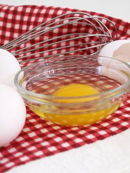 An egg cracked into a dish, eggshells and whole eggs with a whisk laying on a red checkered background.