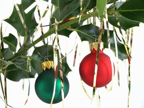 Colorful green and red Christmas bulbs hanging on a branch of holly.  Draped with gold tinsel, over a white background.