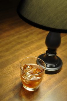 Bourbon on the rocks on wooden table illuminated by lamp.  