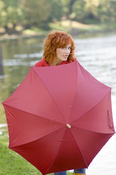 Pretty red haired girl near river playing behind a red umbrella