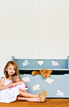 Child playing with her doll while a teddy bear pokes his head out of a toy chest. 